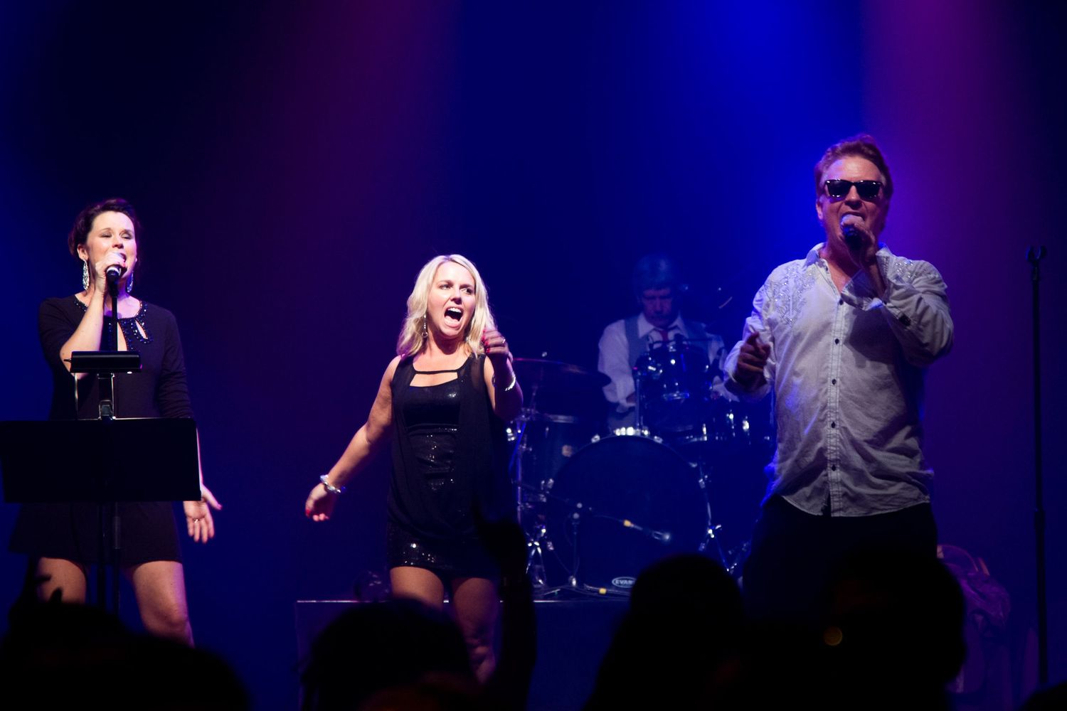 Three singers performing on a lit stage with purple lighting, two women and a man holding microphones at a live concert.