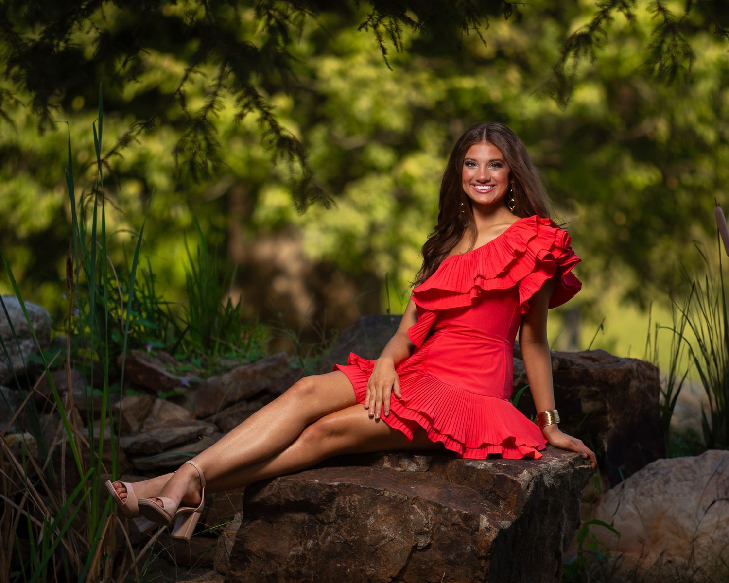 A person in a vibrant red dress poses gracefully on a fallen tree trunk in a lush green forest setting.