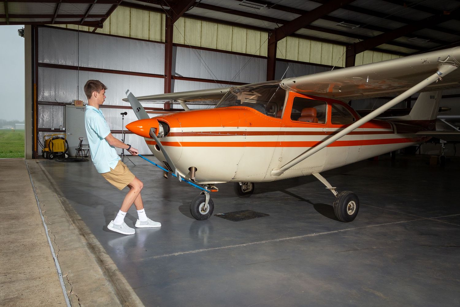 Small orange and white Cessna aircraft parked inside a large aircraft hangar with concrete floor and metal roof structure.