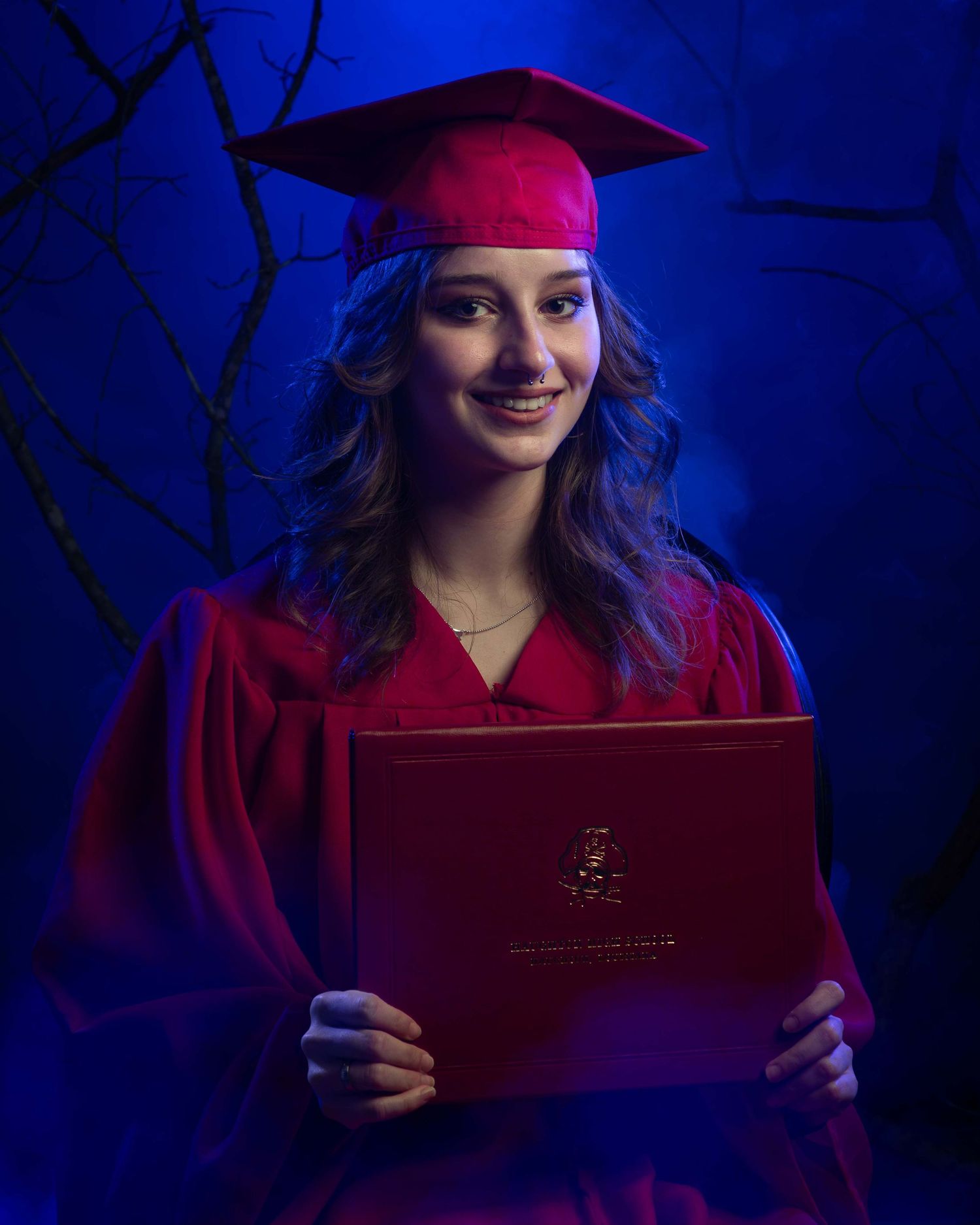 Graduate in red cap and gown holds diploma cover against dramatic blue lighting in professional portrait.