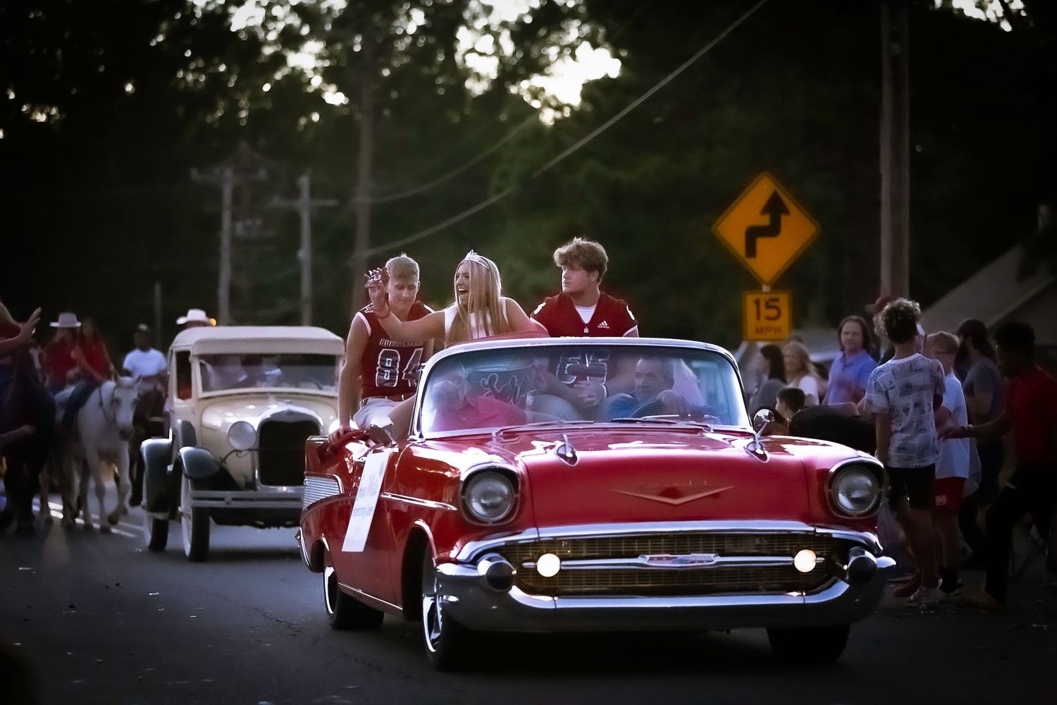 Red 1957 Chevy Bel Air leads a homecoming parade with waving teens, crowds lining the street at dusk.