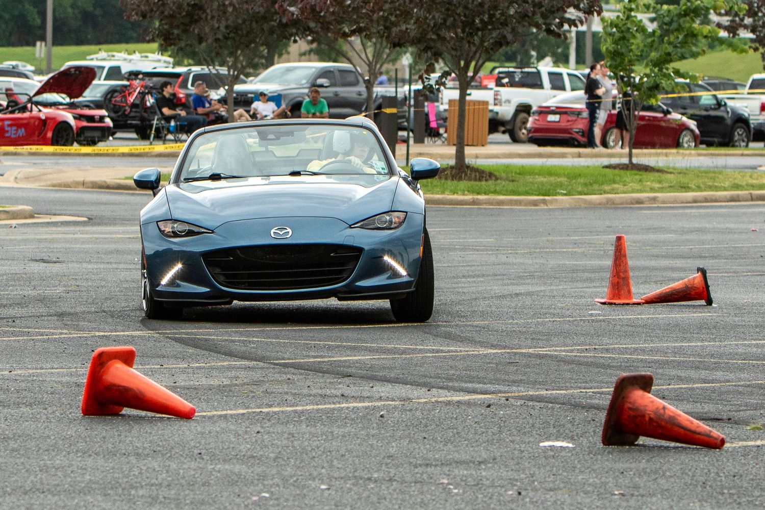 Blue Mazda MX-5 Miata navigating orange cones during an autocross event in a parking lot.