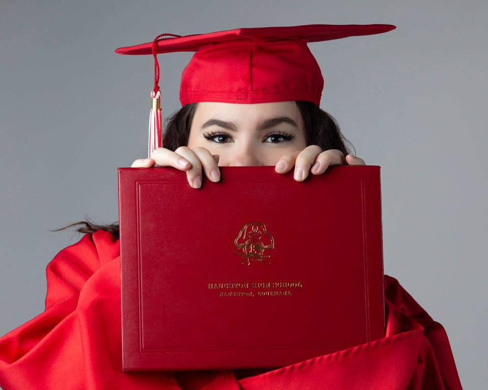 Graduate in bright red cap and gown poses playfully with red diploma holder in creative graduation photo series.