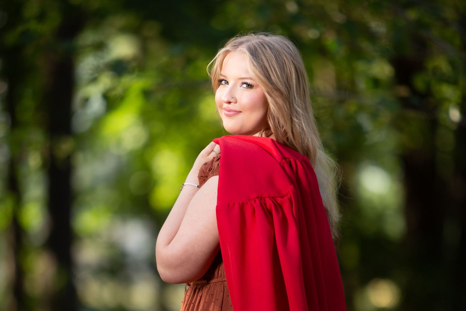 A person wearing a bright red sweater stands in a sunlit outdoor setting with blurred green foliage in the background.