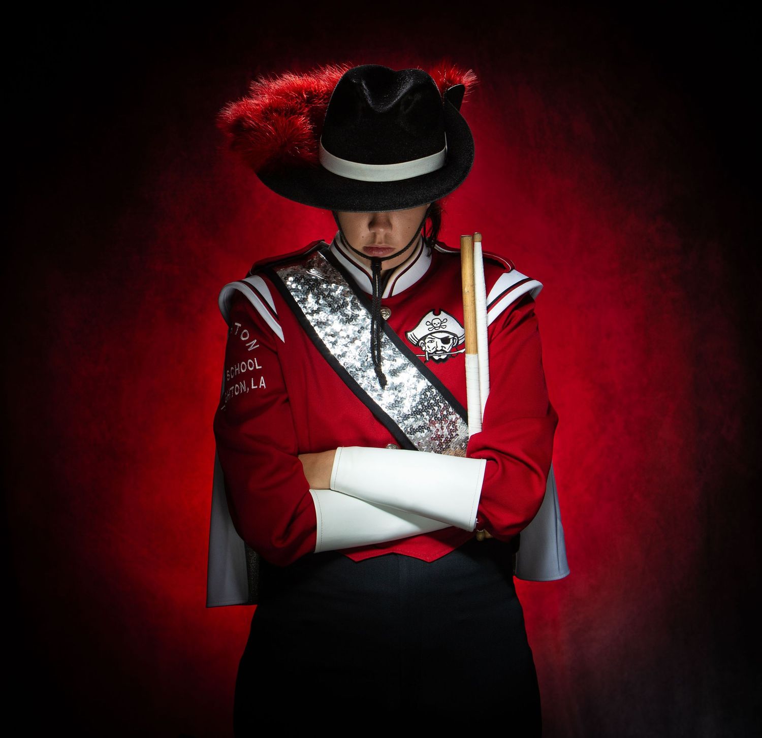 Dramatic portrait of a performer in red and white marching band uniform with black hat against dark red background.
