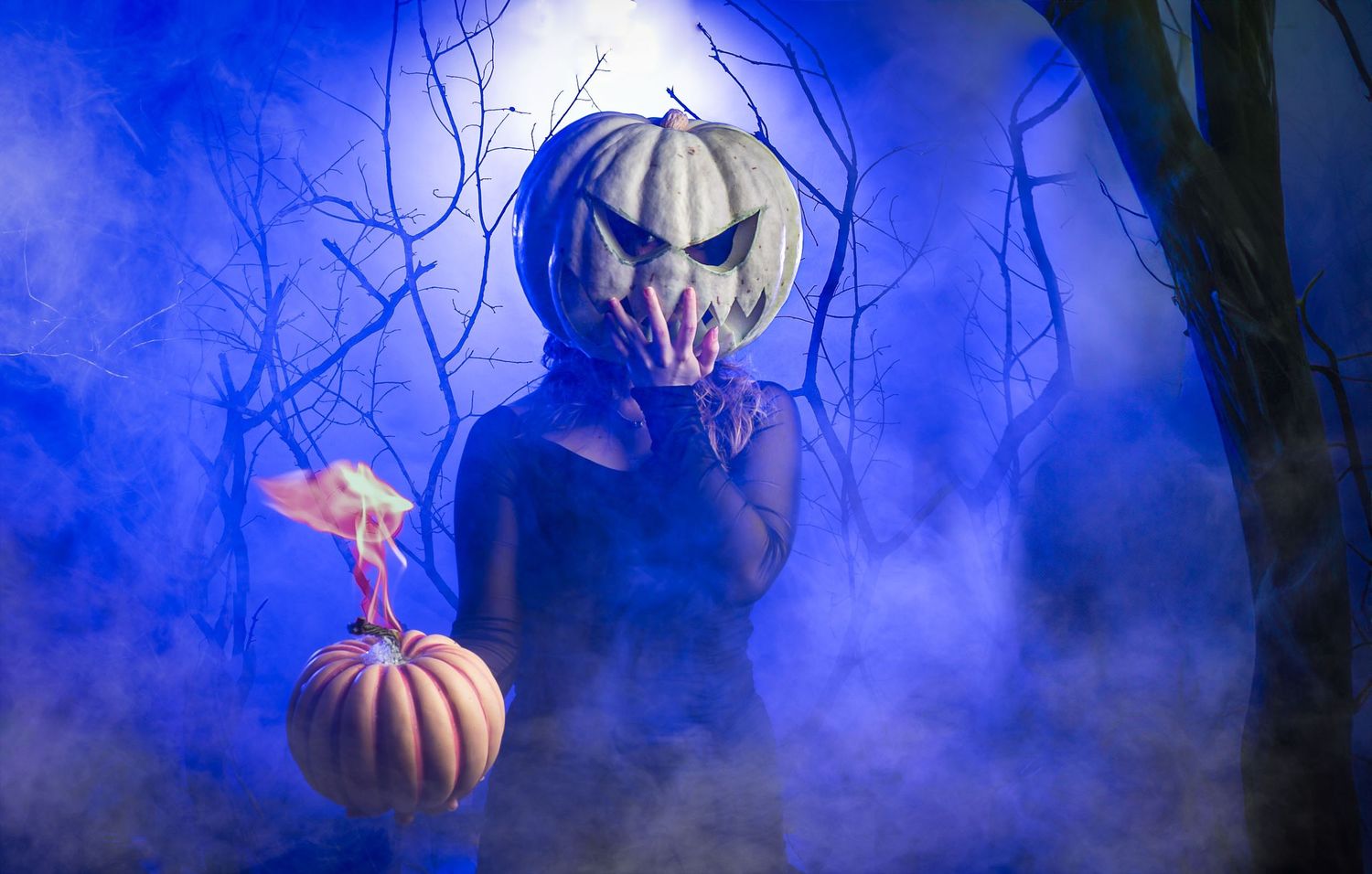 Glowing orange jack o'lanterns float mysteriously in a misty blue forest setting at night.