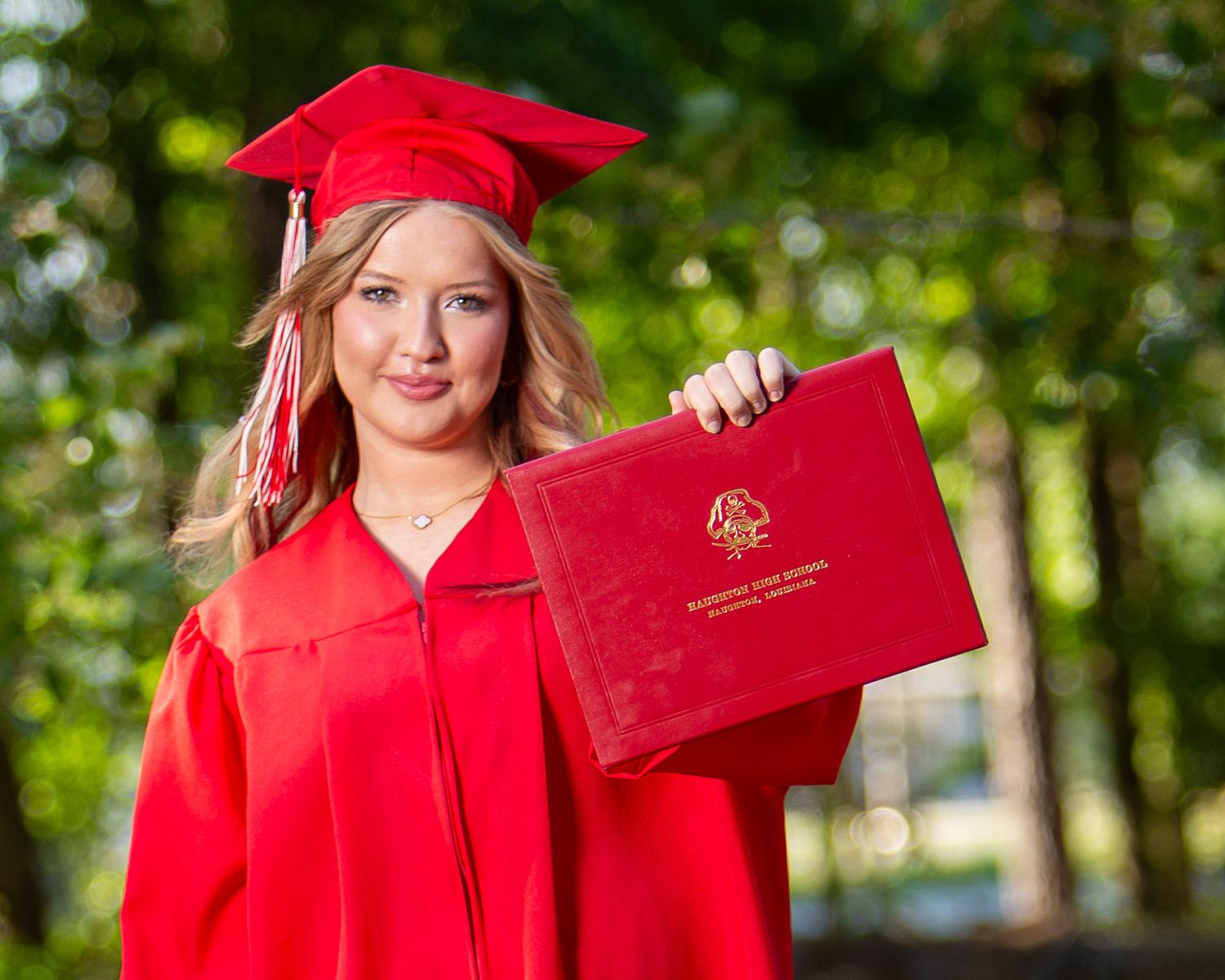 Haughton Louisiana High School Graduate in red cap and gown holds diploma case while posing outdoors against green foliage background.