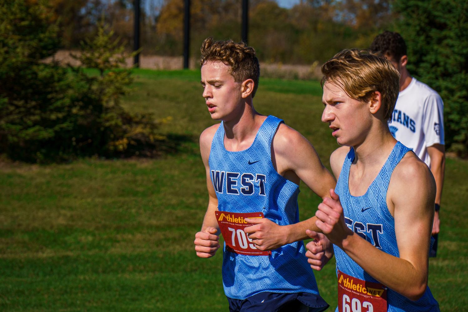 Two high school cross country runners in light blue West jerseys compete during a sunny fall race.