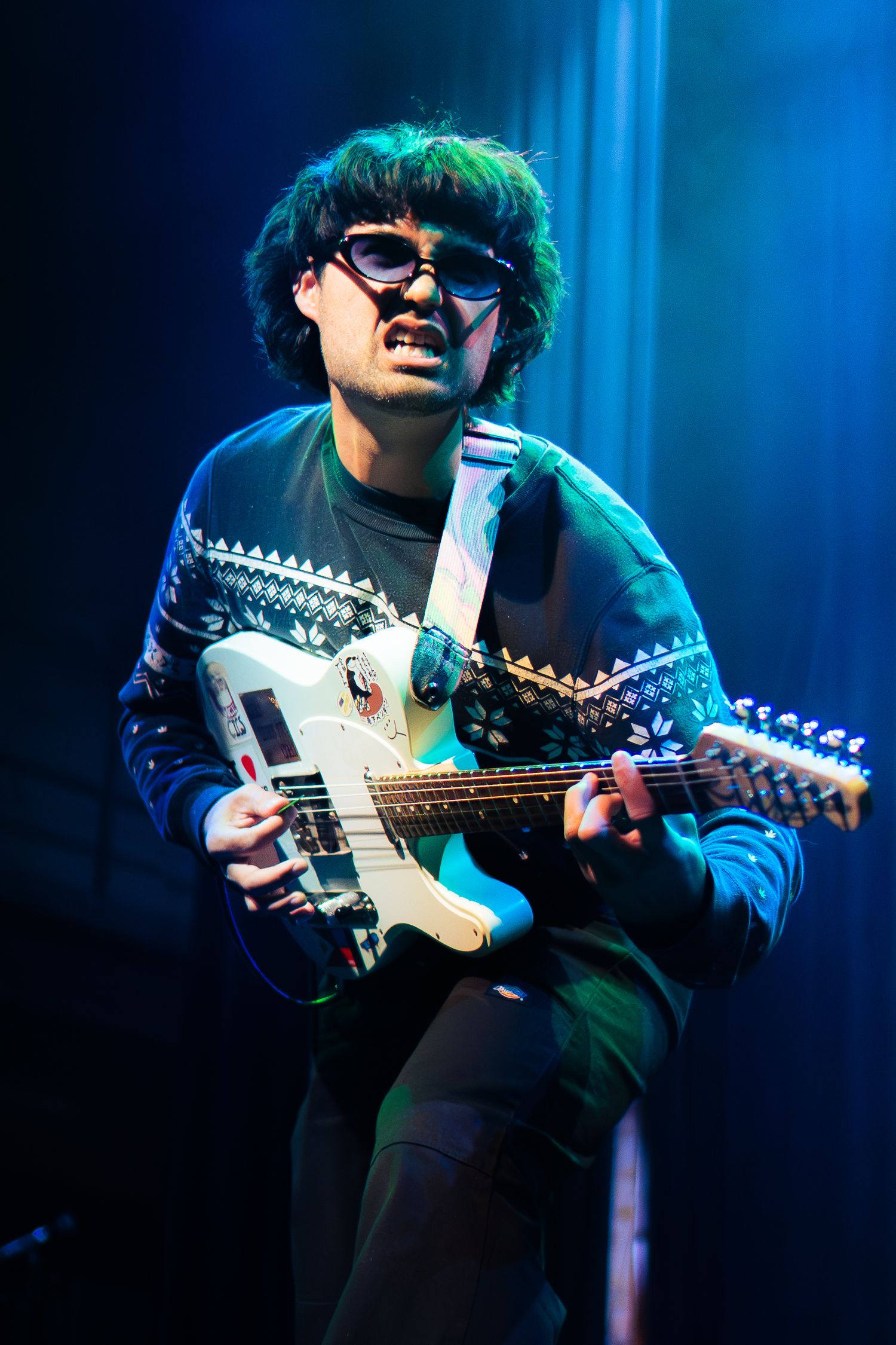 Musician in sunglasses plays electric guitar under dramatic blue stage lighting during a performance.