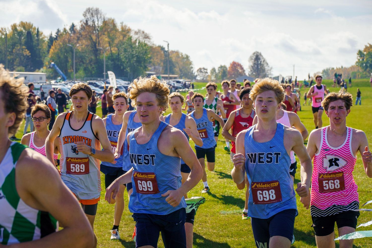 Cross country runners competing in a fall race on a grassy course with spectators cheering from the sidelines.