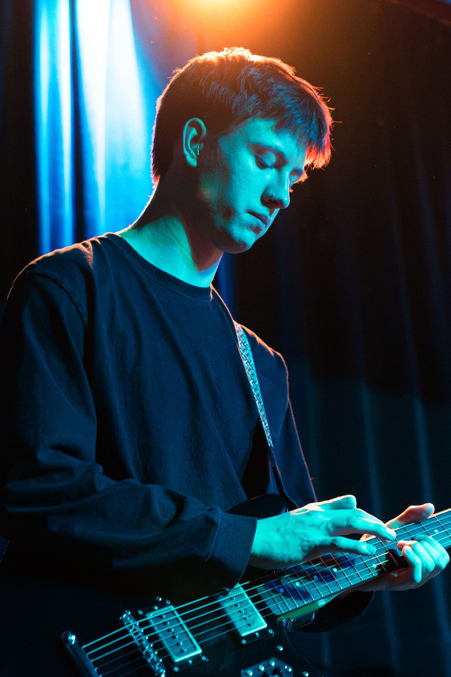 A musician plays an electric bass guitar in dramatic blue and orange stage lighting during a performance.