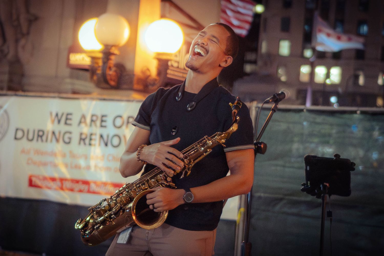 Street musician performs with a saxophone at dusk while illuminated by warm glowing lights in an urban setting.