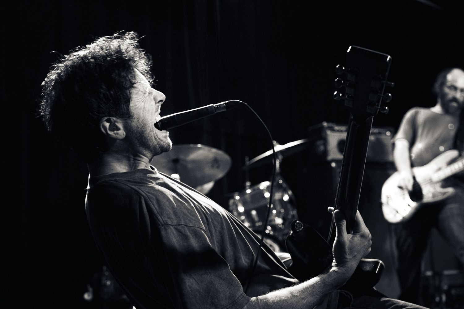 Black and white photo of a male guitarist singing into a microphone on stage with band members performing behind him.