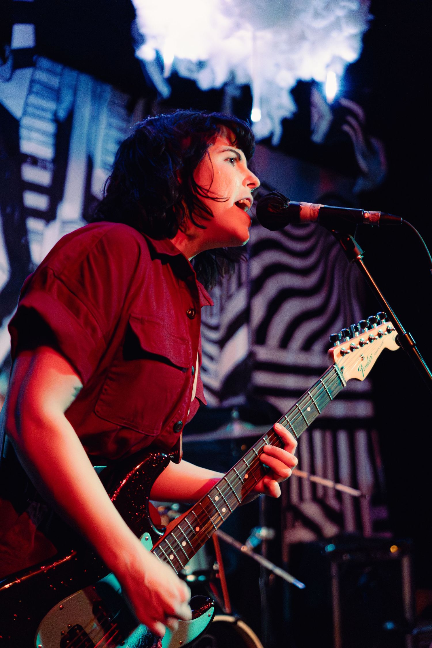 A musician in a red shirt performs energetically with an electric guitar on stage under dramatic lighting.