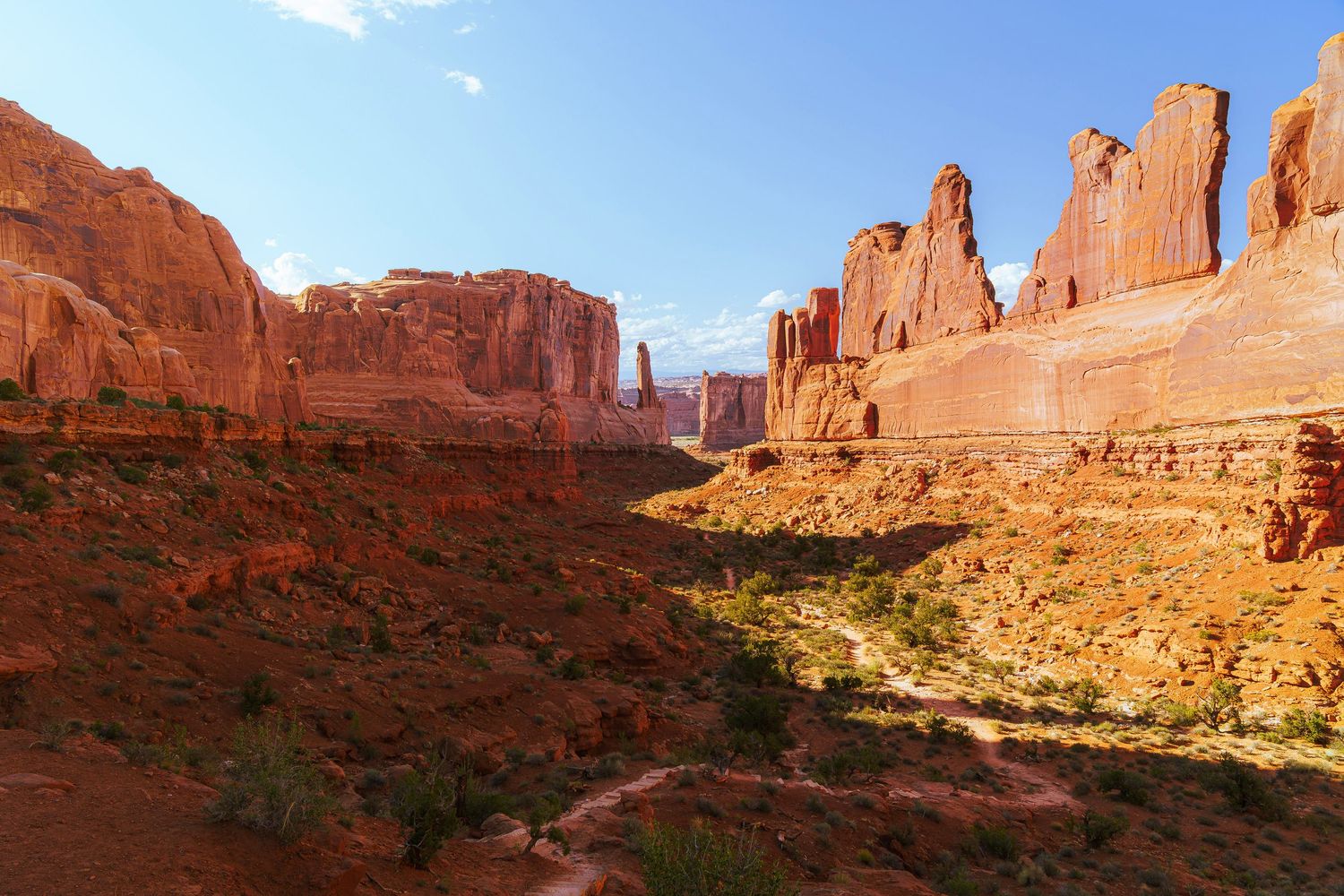 Red sandstone rock formations tower over a scenic desert canyon under a clear blue sky in Arches National Park, Utah.