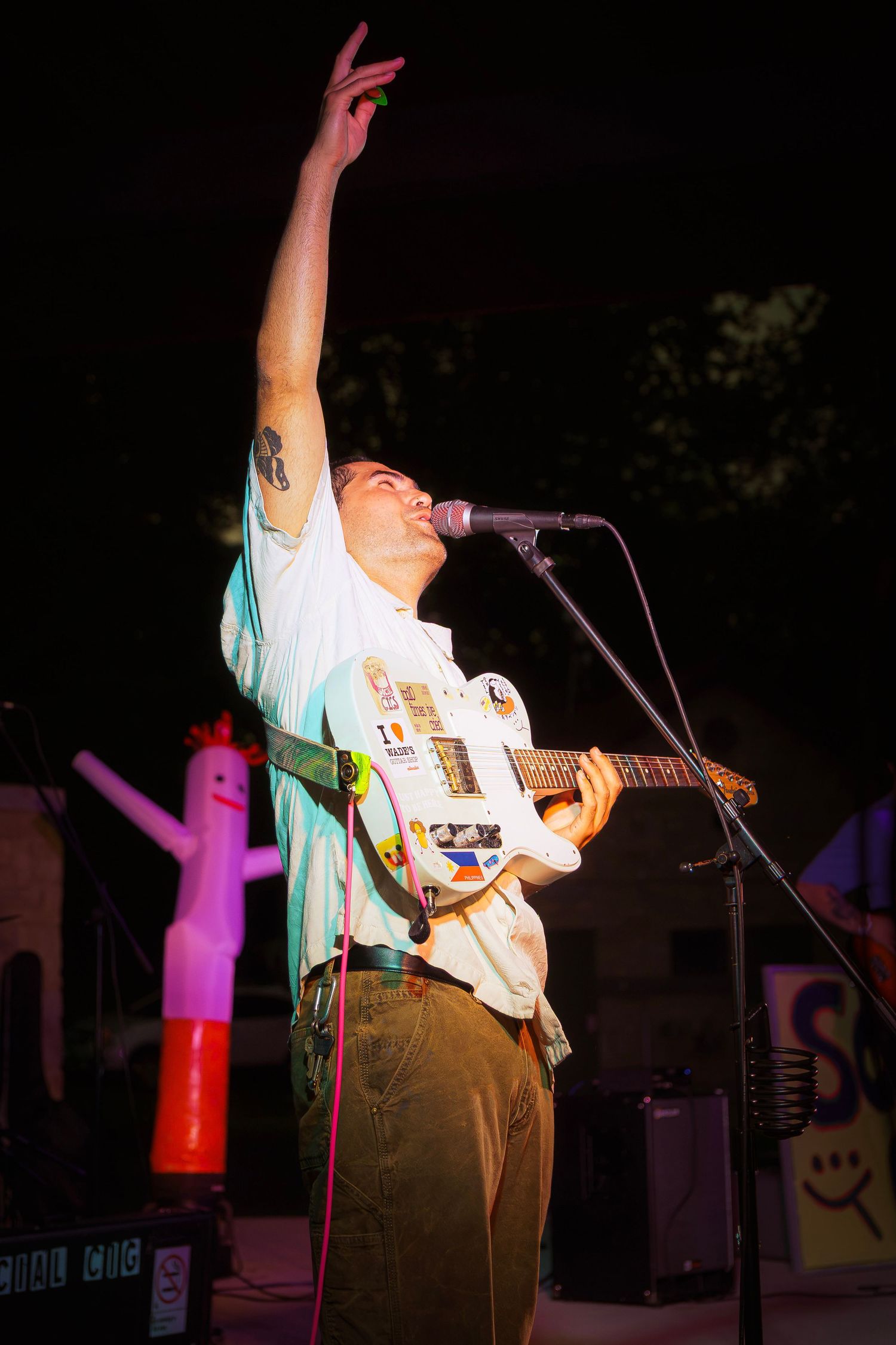 Male guitarist performing live on stage, raising one hand with sticker-covered electric guitar at outdoor night concert.