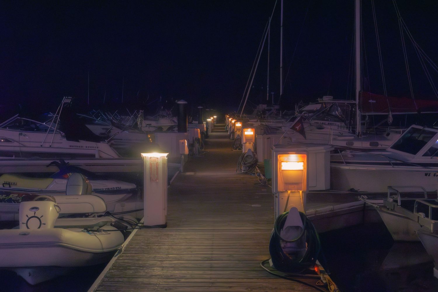 Boats docked at a marina at dusk with glowing lights reflecting on the calm water.