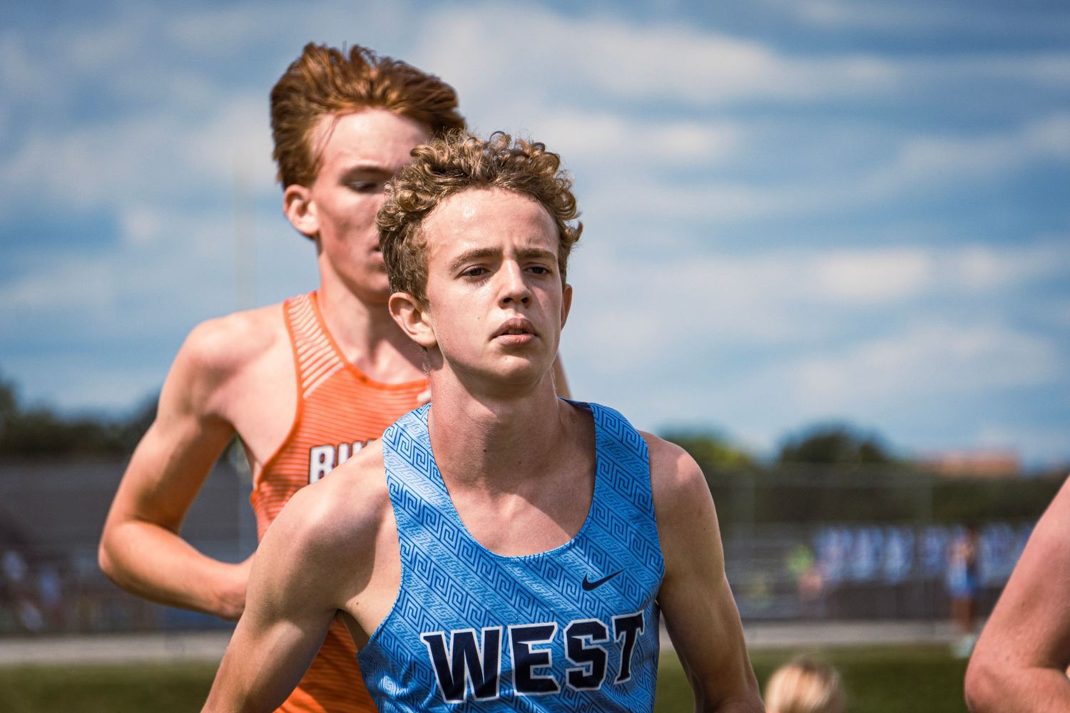 Runners wearing race bibs compete in a cross country meet under blue skies.