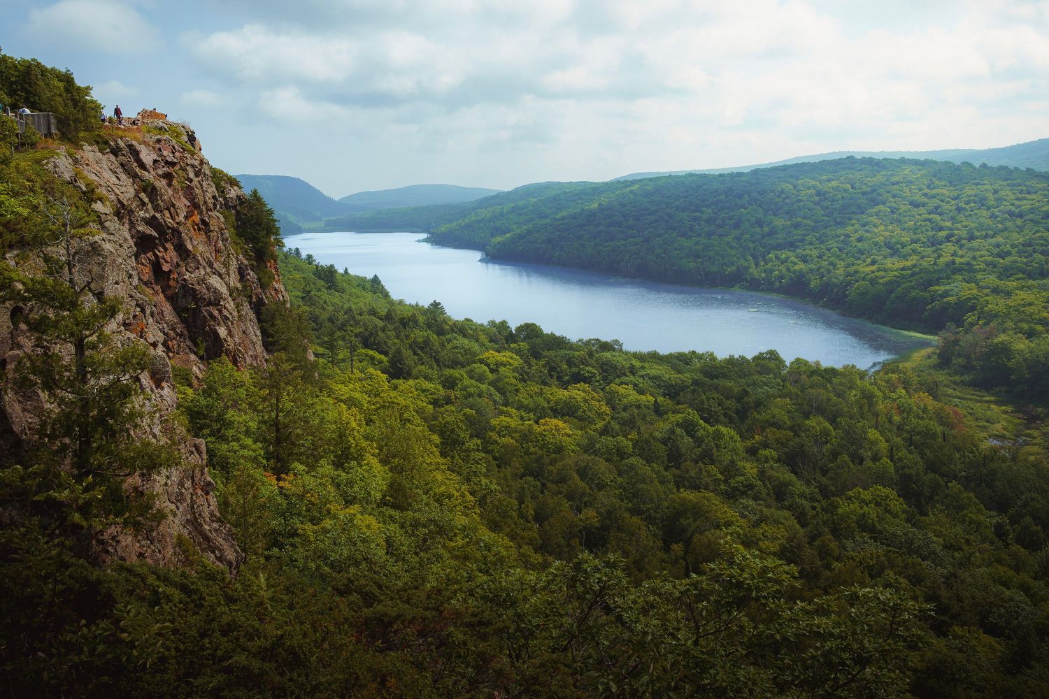 Panoramic view of a scenic lake surrounded by lush green mountains and rocky cliffs on a sunny summer day.