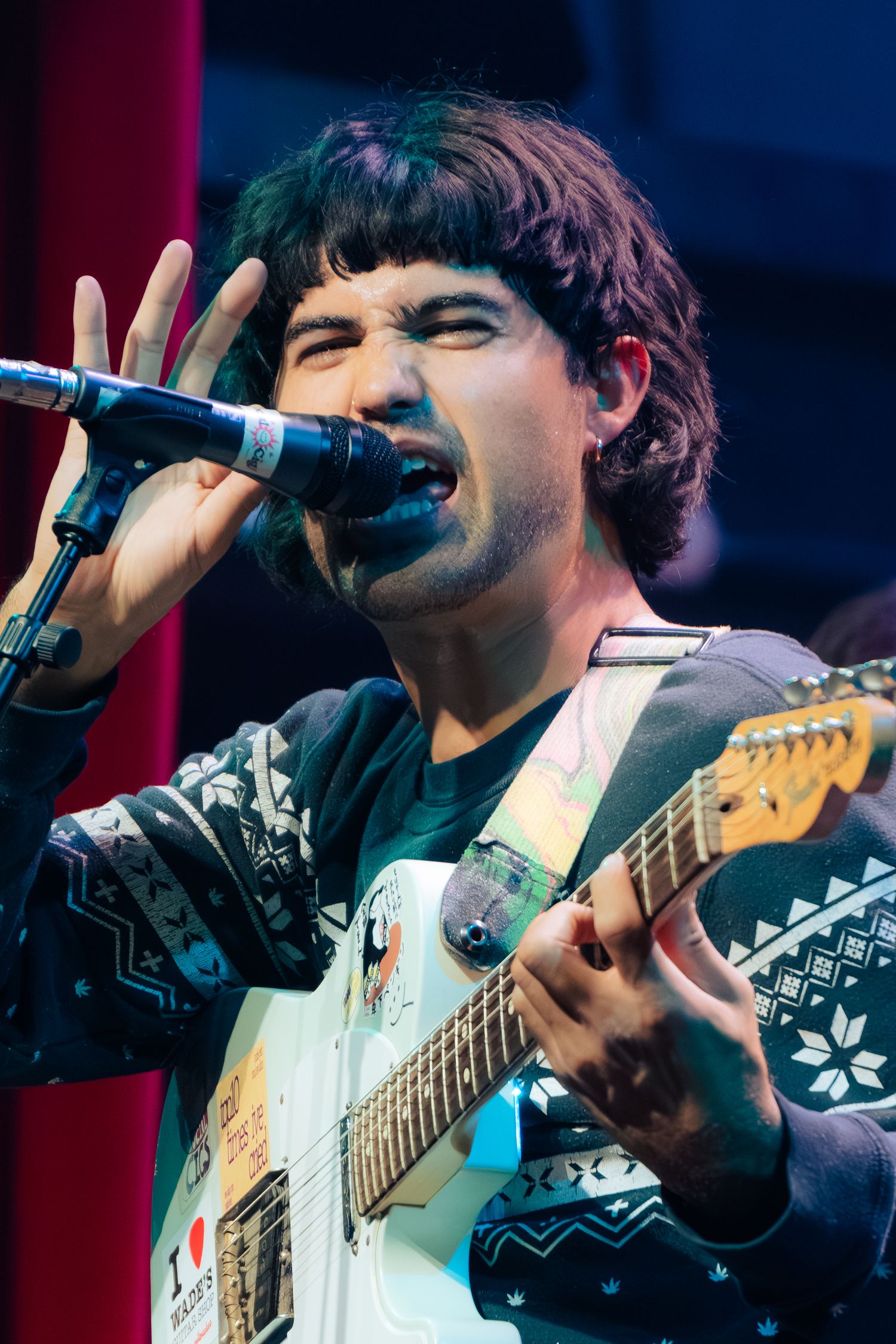 Musician performs on stage wearing a winter-themed sweater while playing electric guitar and singing into microphone.