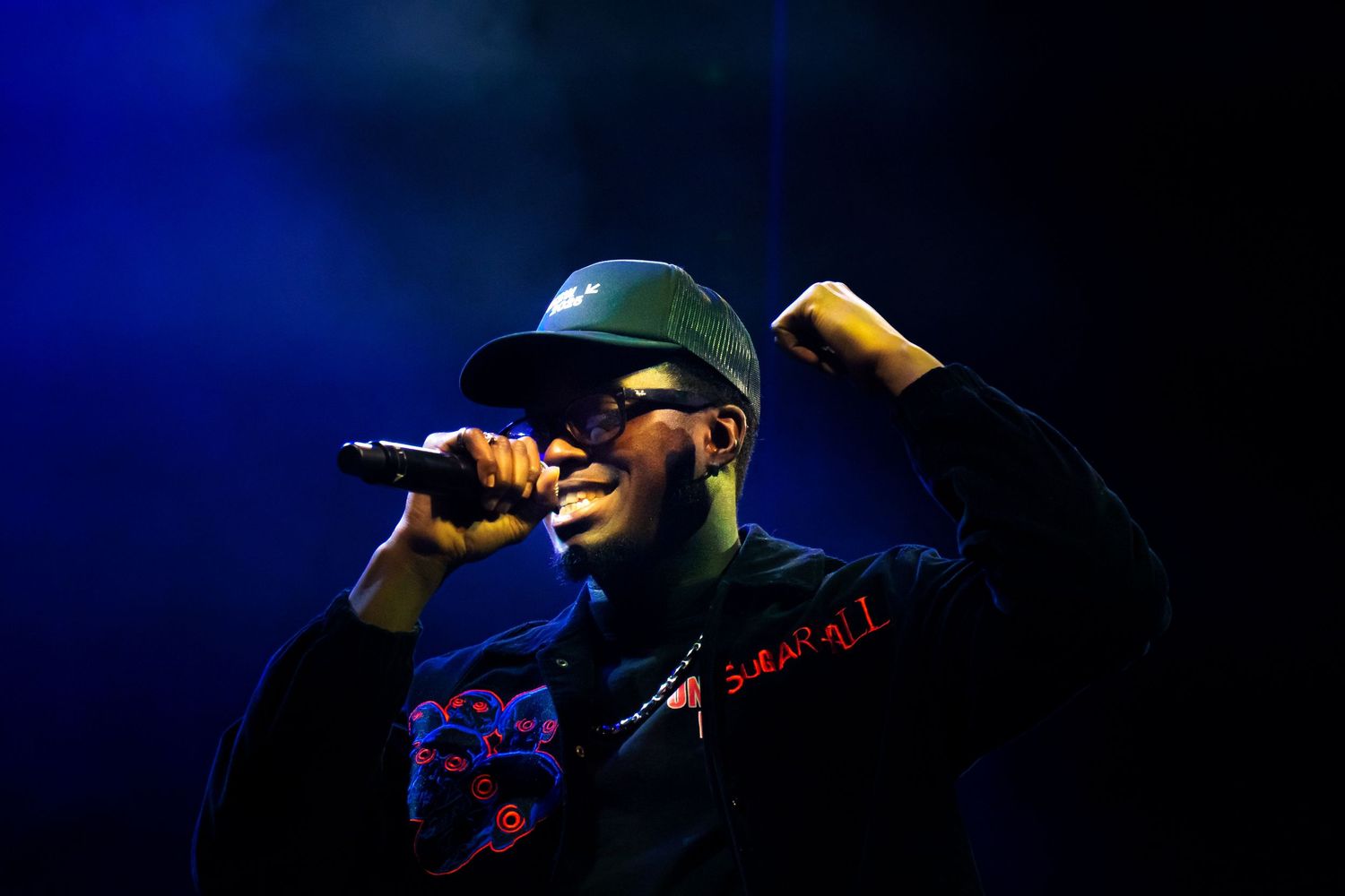 A performer holds a microphone on stage during a concert with dramatic blue lighting in the background.