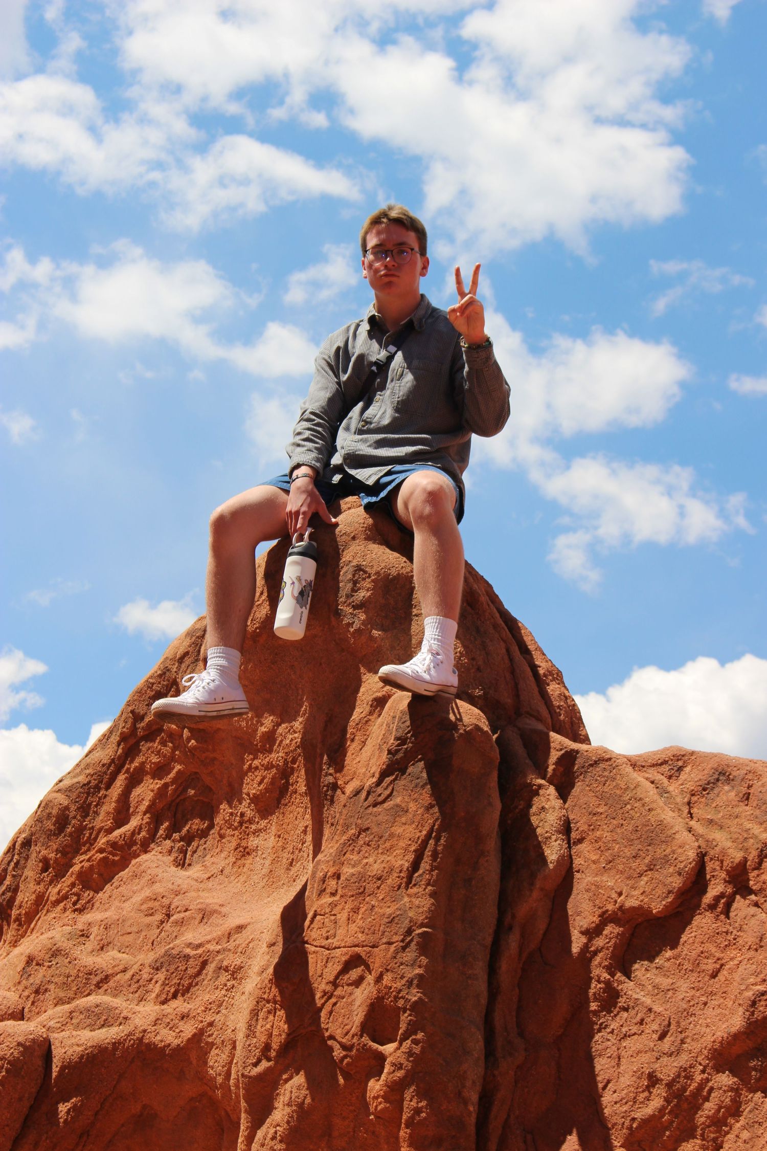 Someone sits atop a red rock formation against a bright blue sky with white puffy clouds.