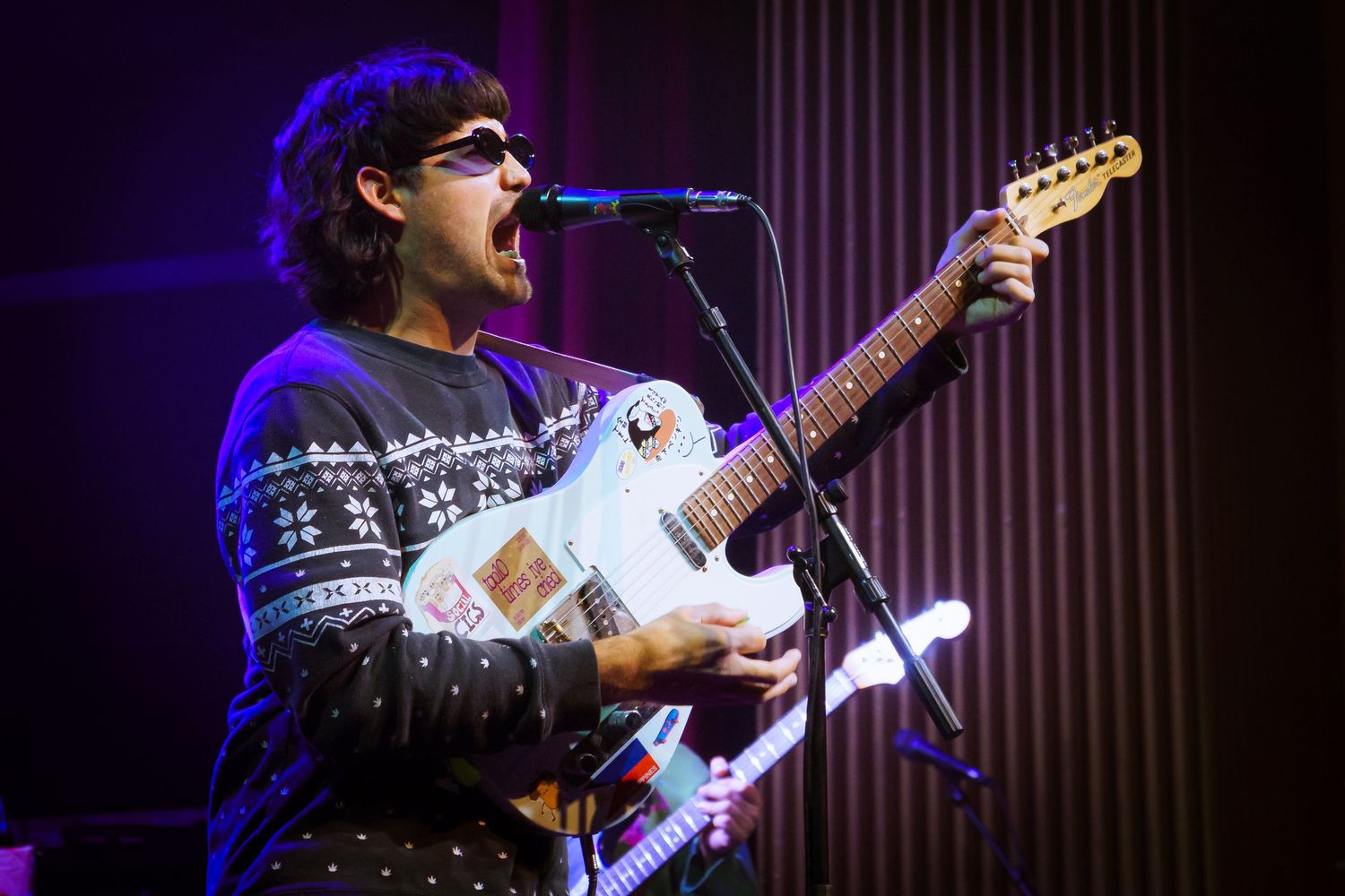 A musician performs on stage with a white electric guitar under blue stage lighting.