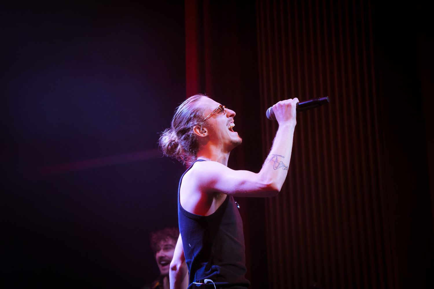 A performer in a black tank top sings passionately on stage under dramatic purple lighting during a concert performance.