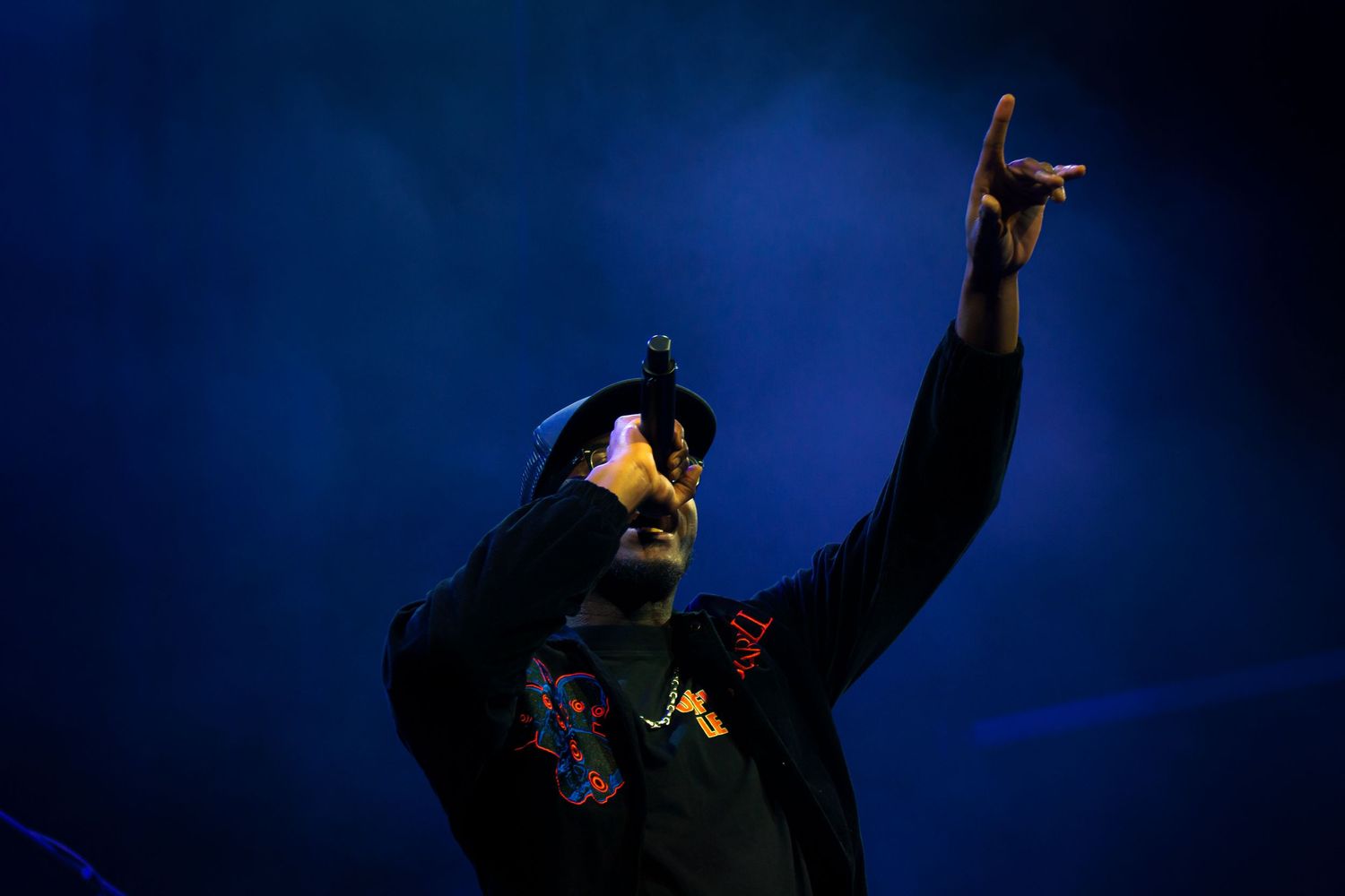 A performer in black clothing raises their hand against a dark blue background during a stage performance.