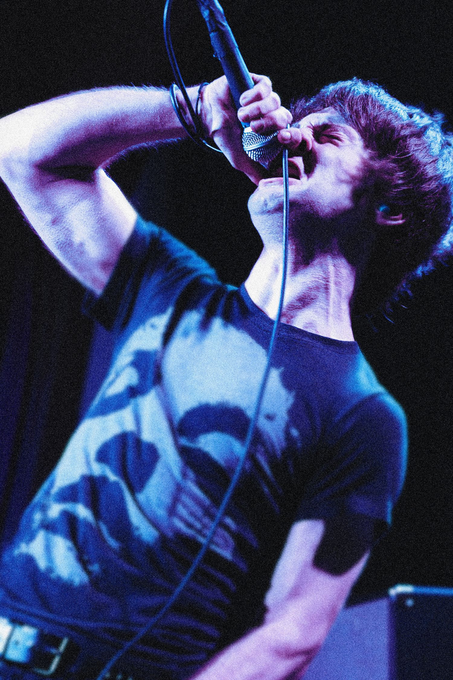 A performer in a dark t-shirt sings passionately into a microphone on stage under dramatic blue lighting.