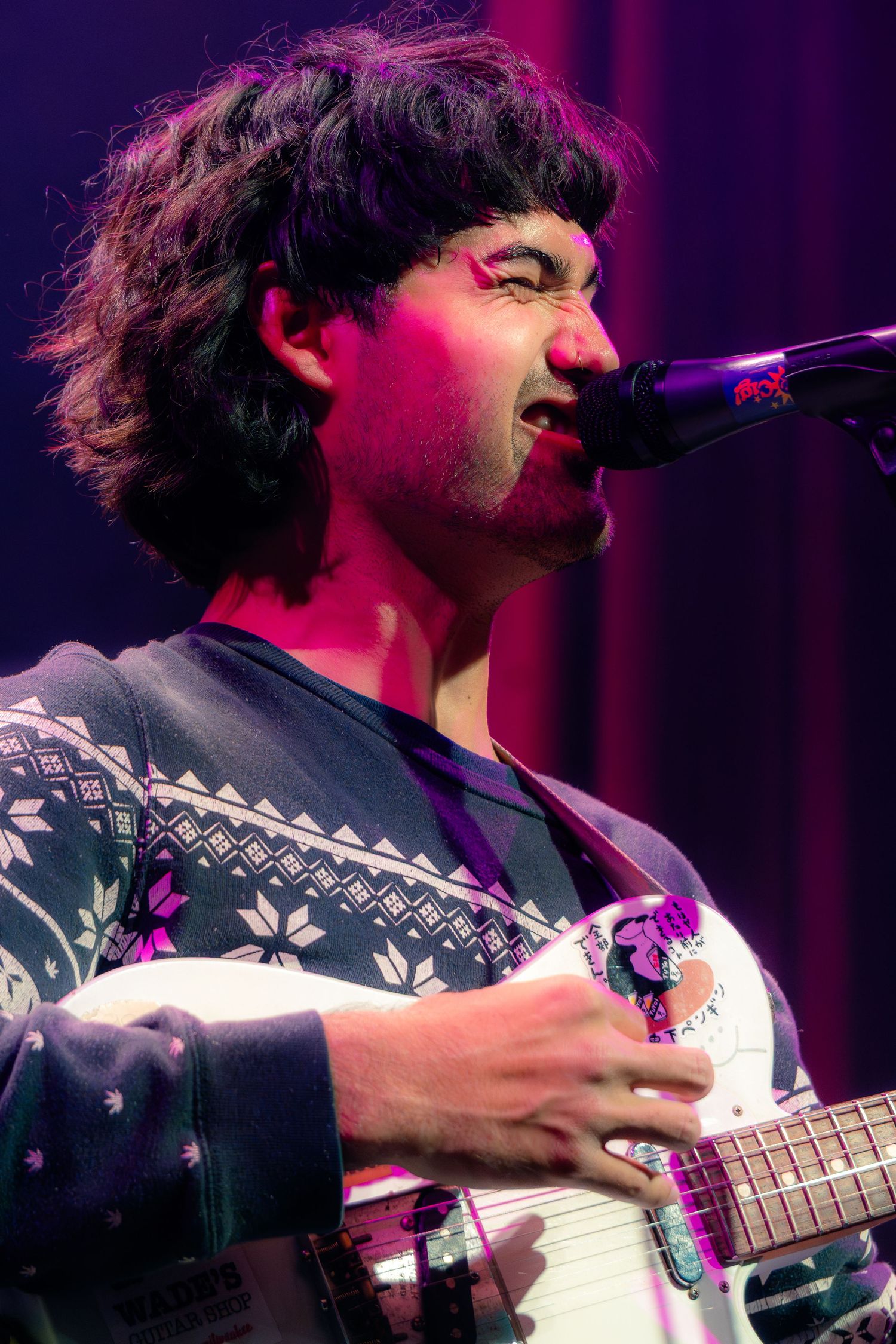 A musician performs on stage wearing a patterned sweater while playing guitar under purple lighting.