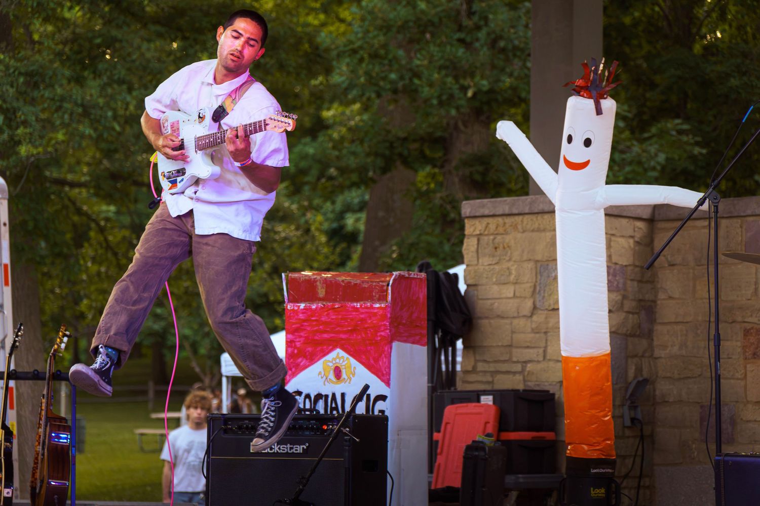 Guitarist jumps on outdoor stage beside inflatable cigarette costume mascot during live music performance.