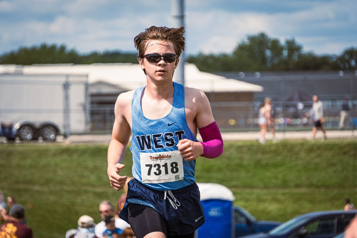 Runners compete in an outdoor track and field event on a sunny day with spectators watching from the sidelines.