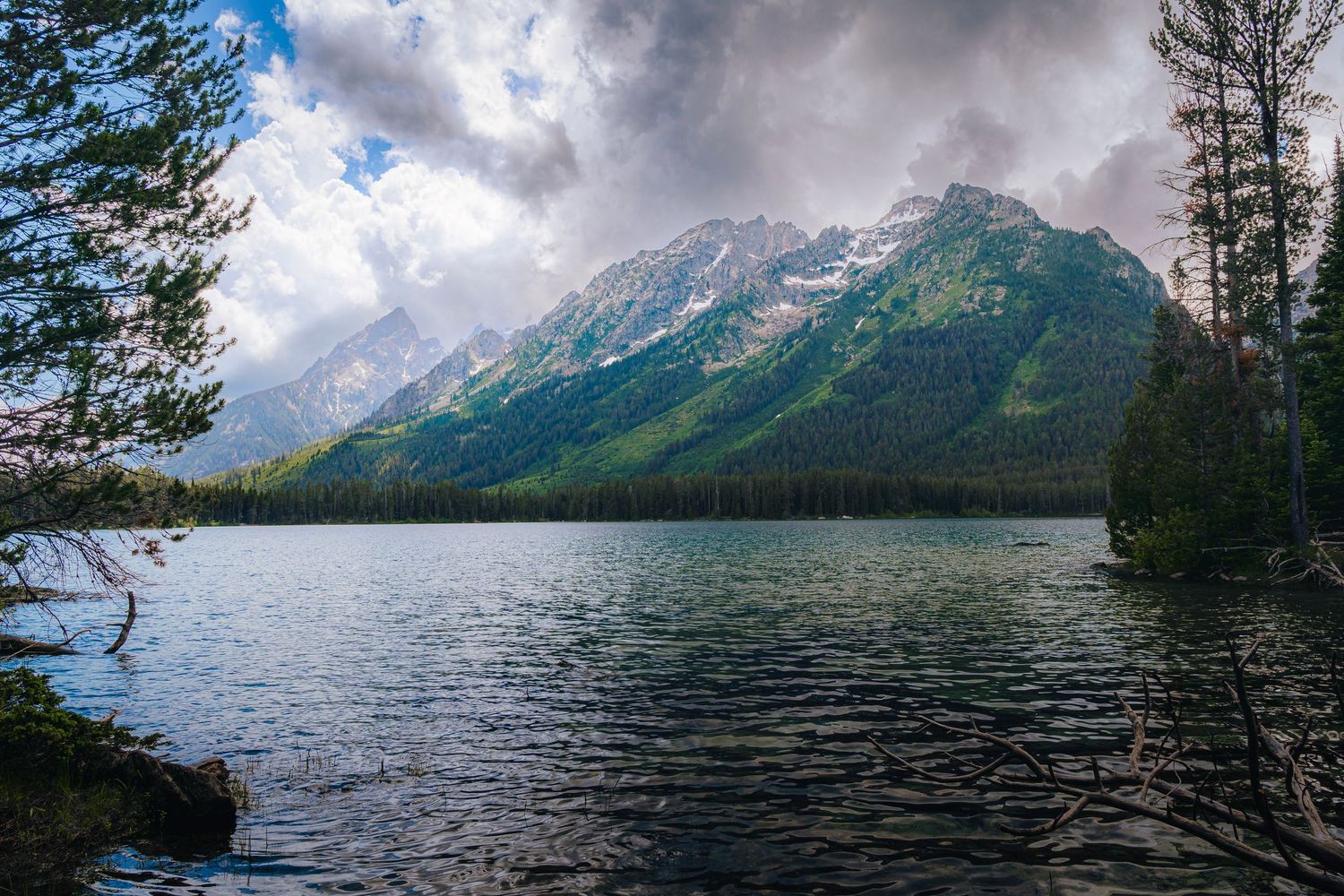 Panoramic view of a mountain lake surrounded by evergreen trees with dramatic cloudy skies above.