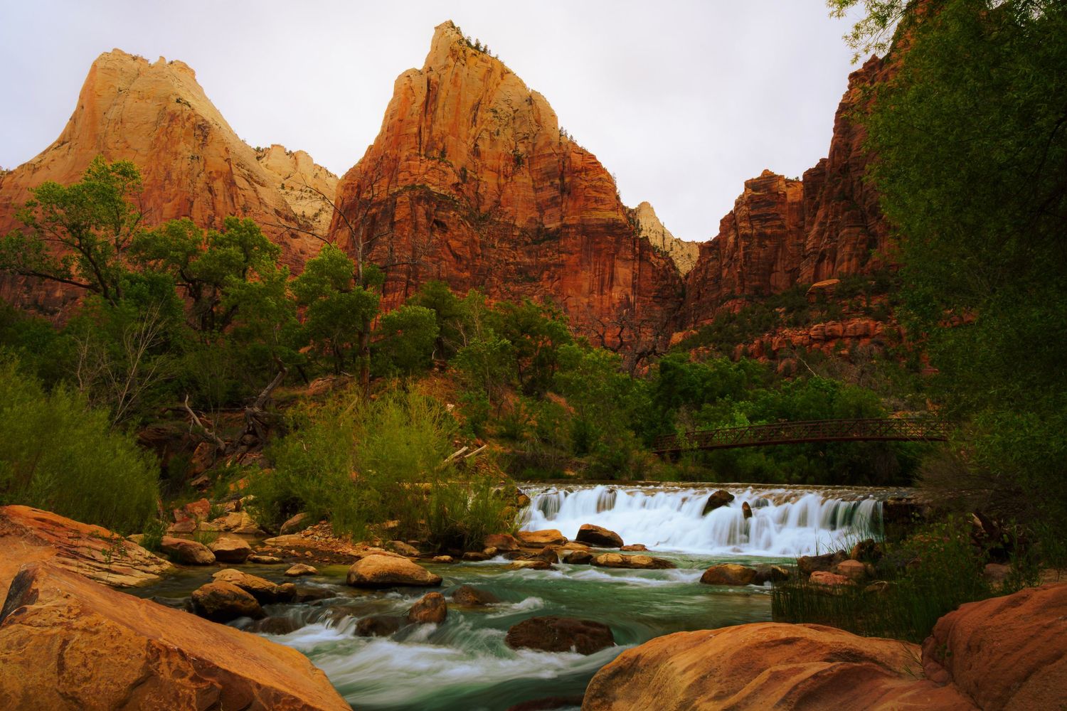 Scenic Zion National Park waterfall with red rock formations, lush green trees, and rushing blue water over rocky terrain.