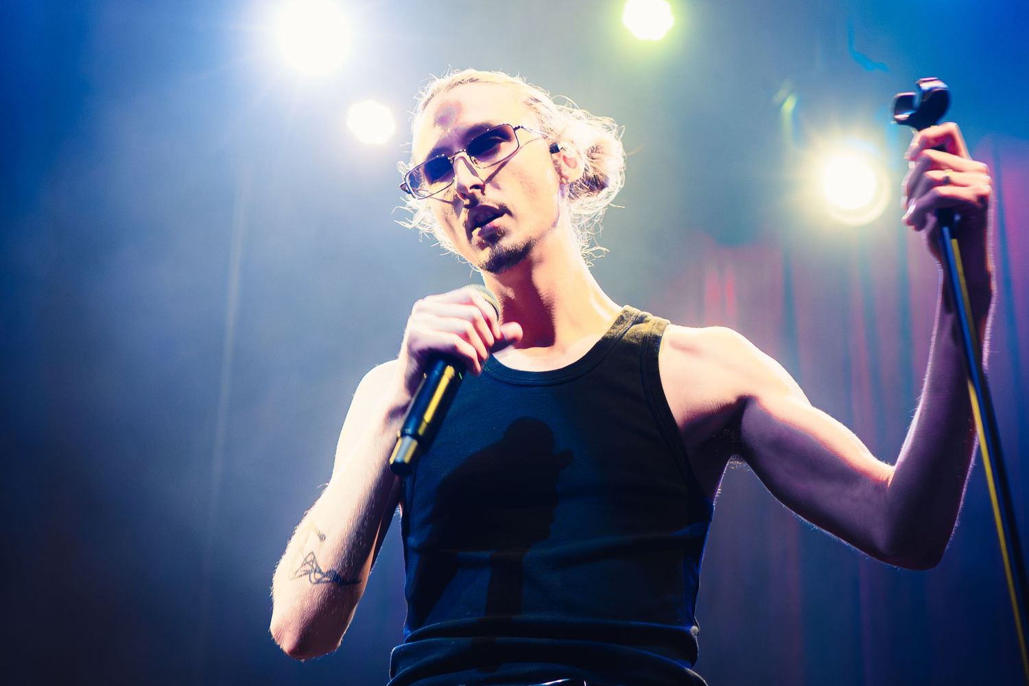 A performer in a black tank top sings passionately on stage with dramatic blue lighting and spotlights in the background.