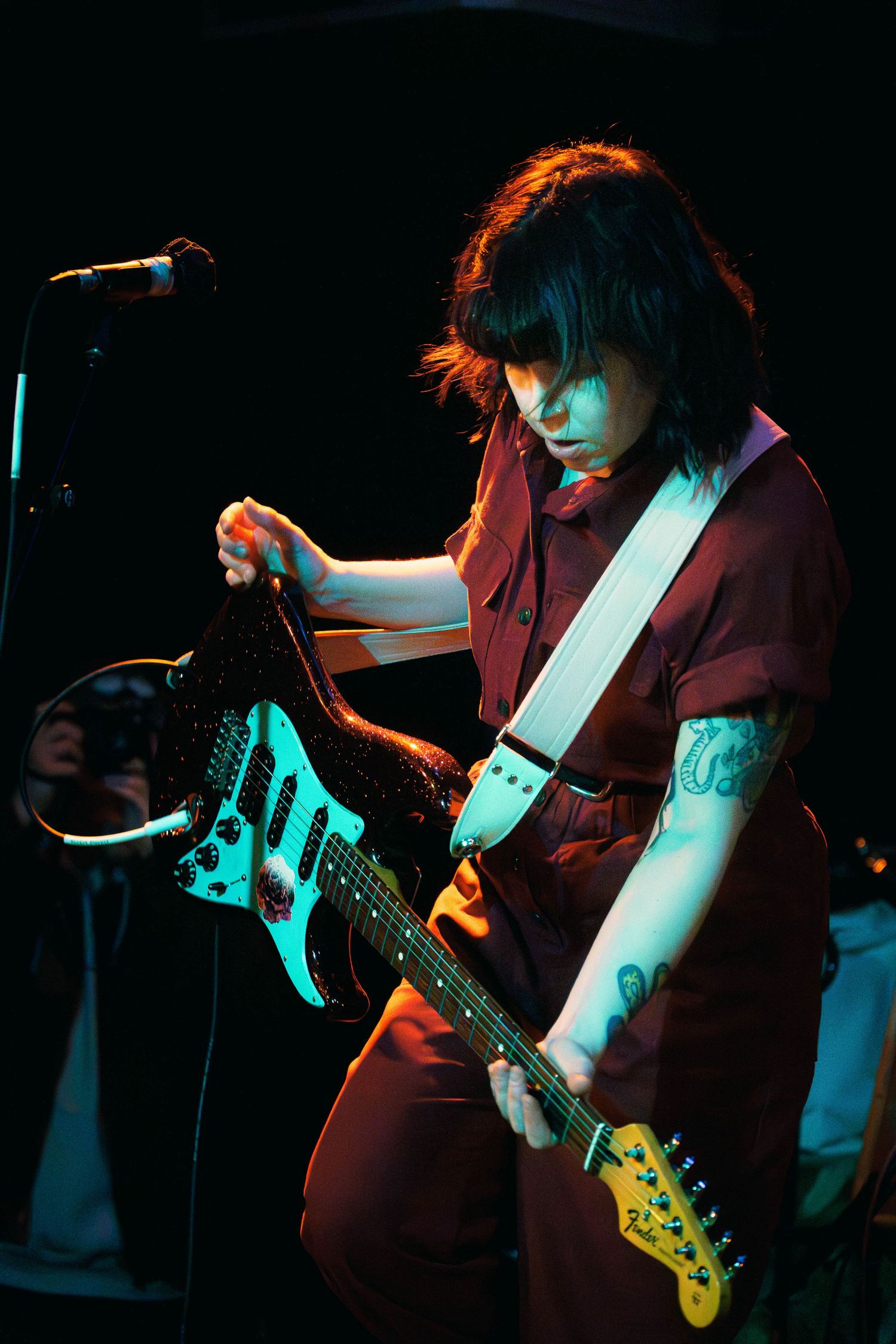 A guitarist in a dark red outfit performs on stage with a white electric guitar under dramatic blue lighting.