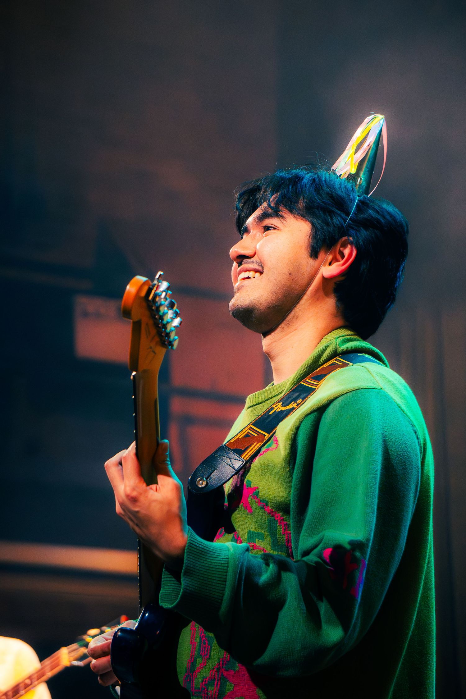 Musician in green hoodie performs passionately on stage with dramatic lighting and expressions while playing guitar.