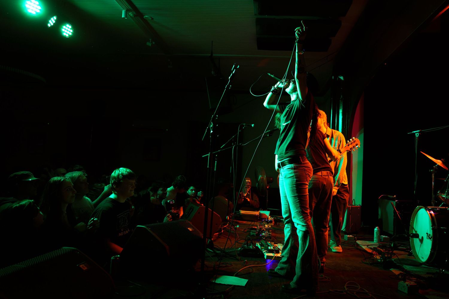 Silhouetted performers on a dark stage illuminated by dramatic green and orange theatrical lighting.