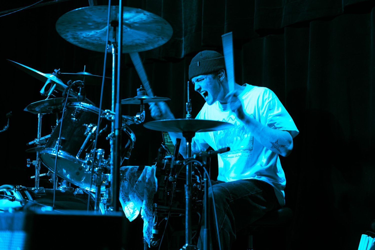 A drummer performs on stage illuminated by dramatic blue lighting during a live music performance.