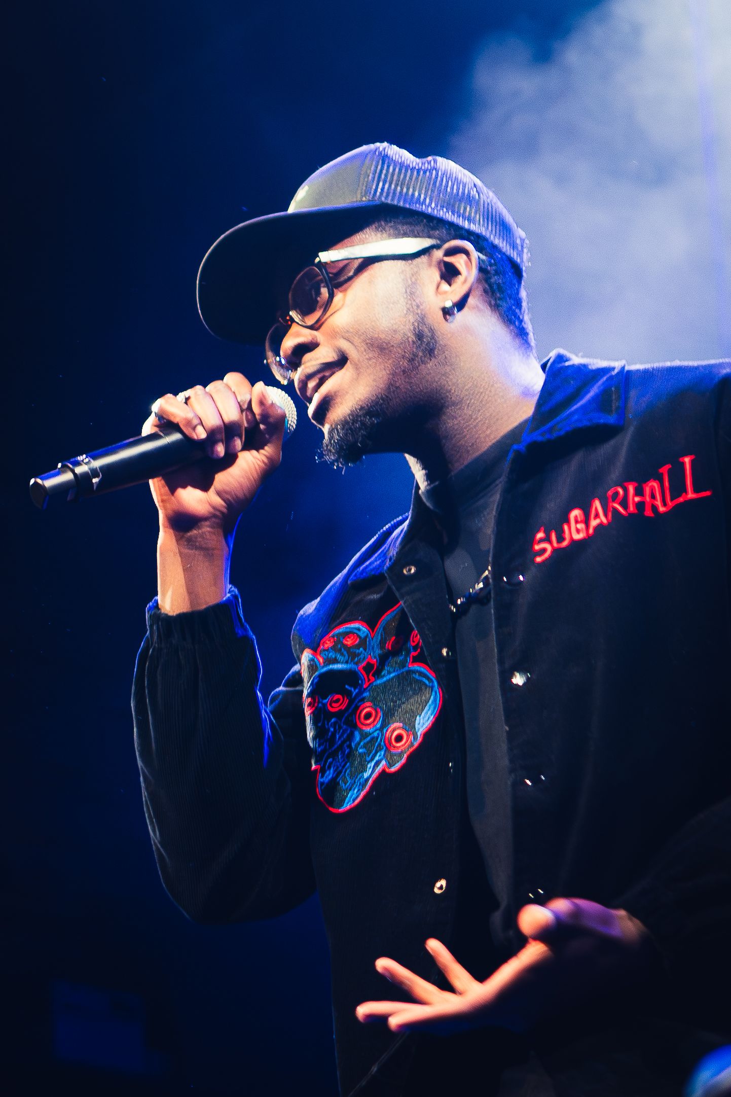 Hip hop performer in dark clothing and baseball cap sings into microphone against blue-lit stage backdrop.