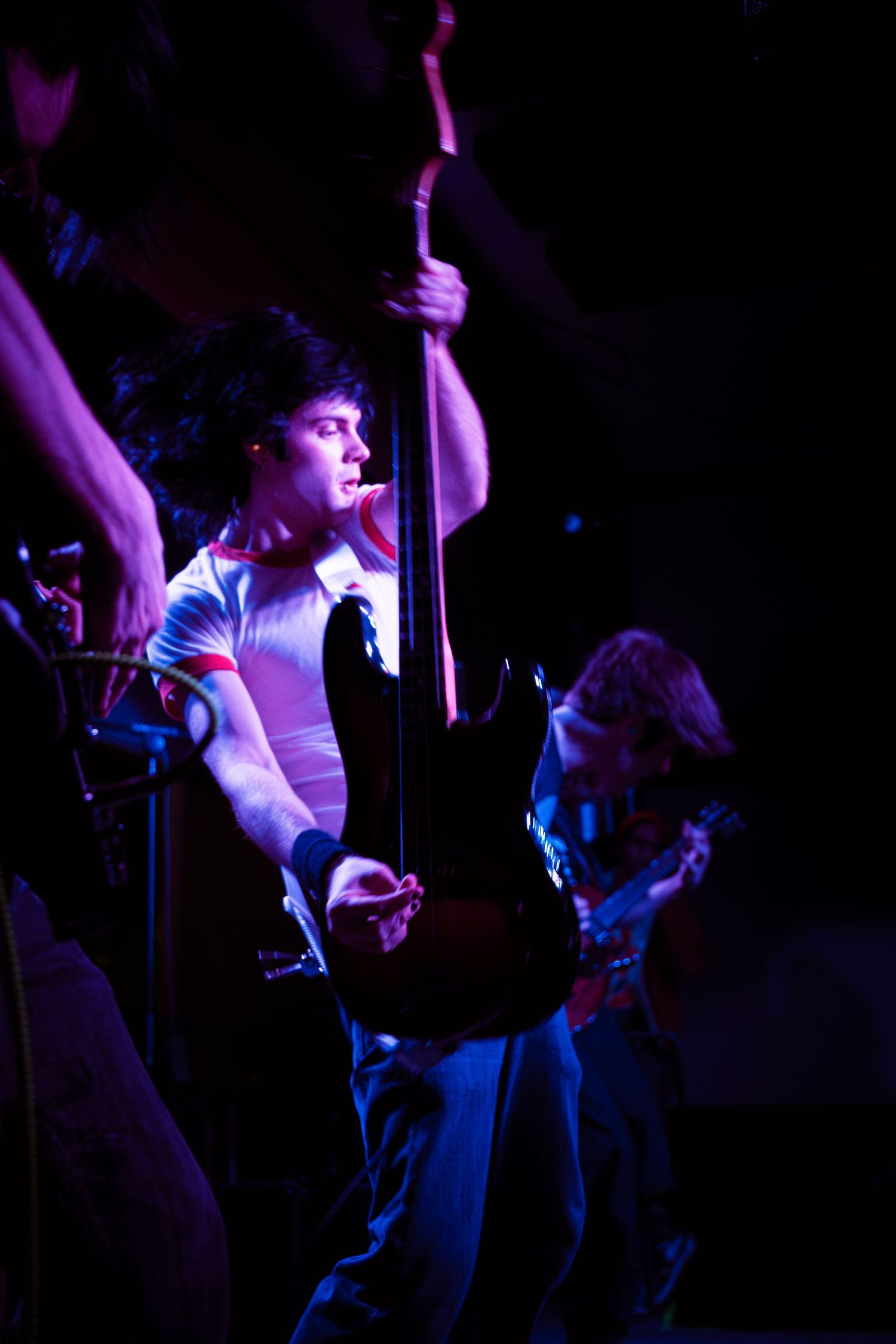 Performer in a white shirt energetically singing on a dimly lit stage with dramatic purple and blue lighting effects.