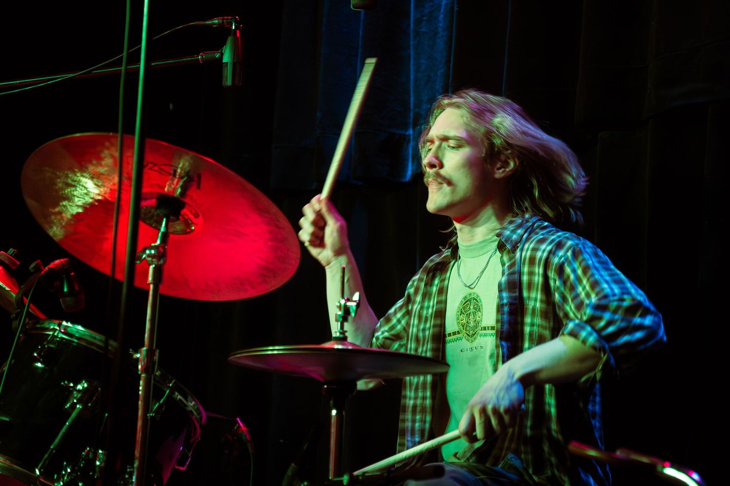 A drummer performs energetically on stage with red and blue lighting while playing a drum kit during a live performance.