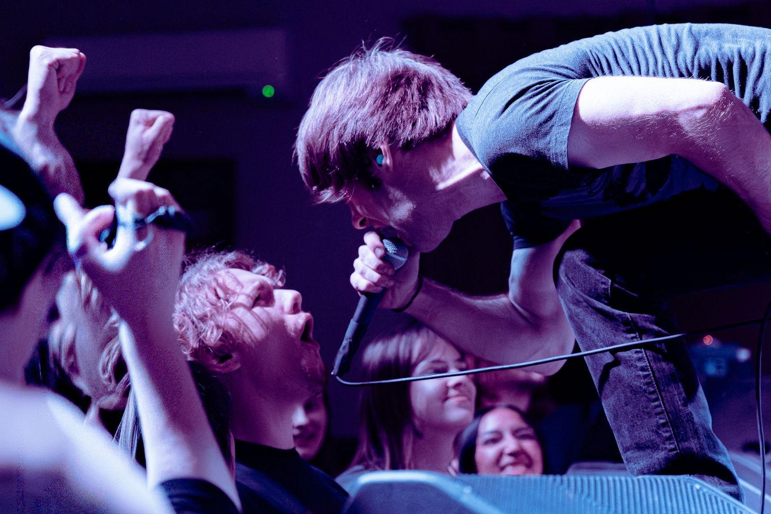 Concert crowd energetically dances and jumps with hands raised at a live music performance in purple lighting.