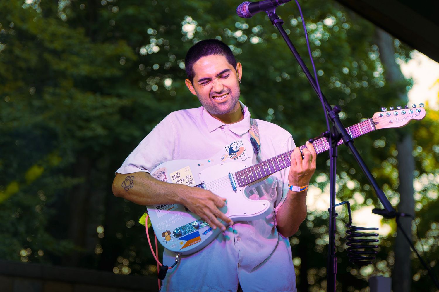 Smiling male guitarist playing white Stratocaster electric guitar on outdoor stage at live music event.