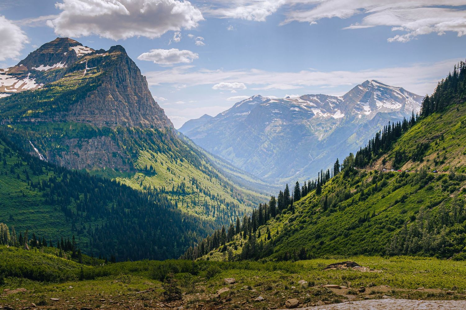 Panoramic view of snow-capped mountains and green valleys in Glacier National Park under a blue cloudy sky.
