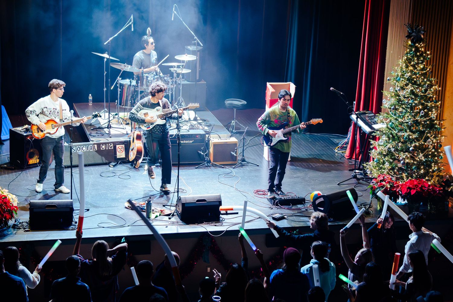 Musicians perform on stage during a live concert with instruments and holiday decorations visible in the background.