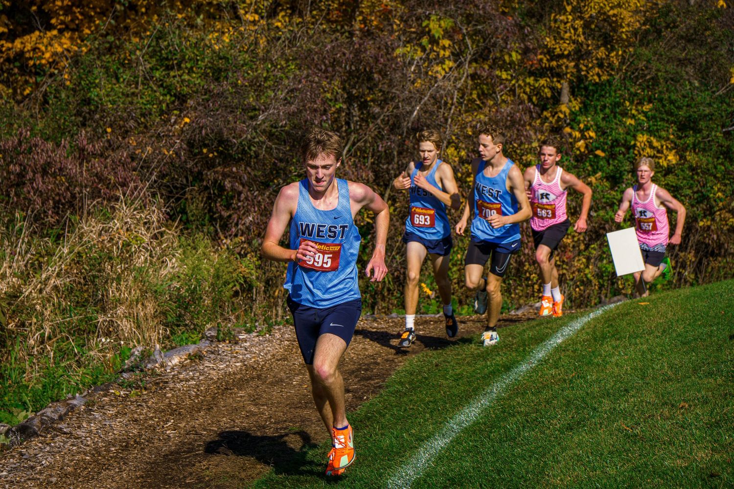 Cross country runners compete on a grassy trail during a fall race with autumn foliage in the background.