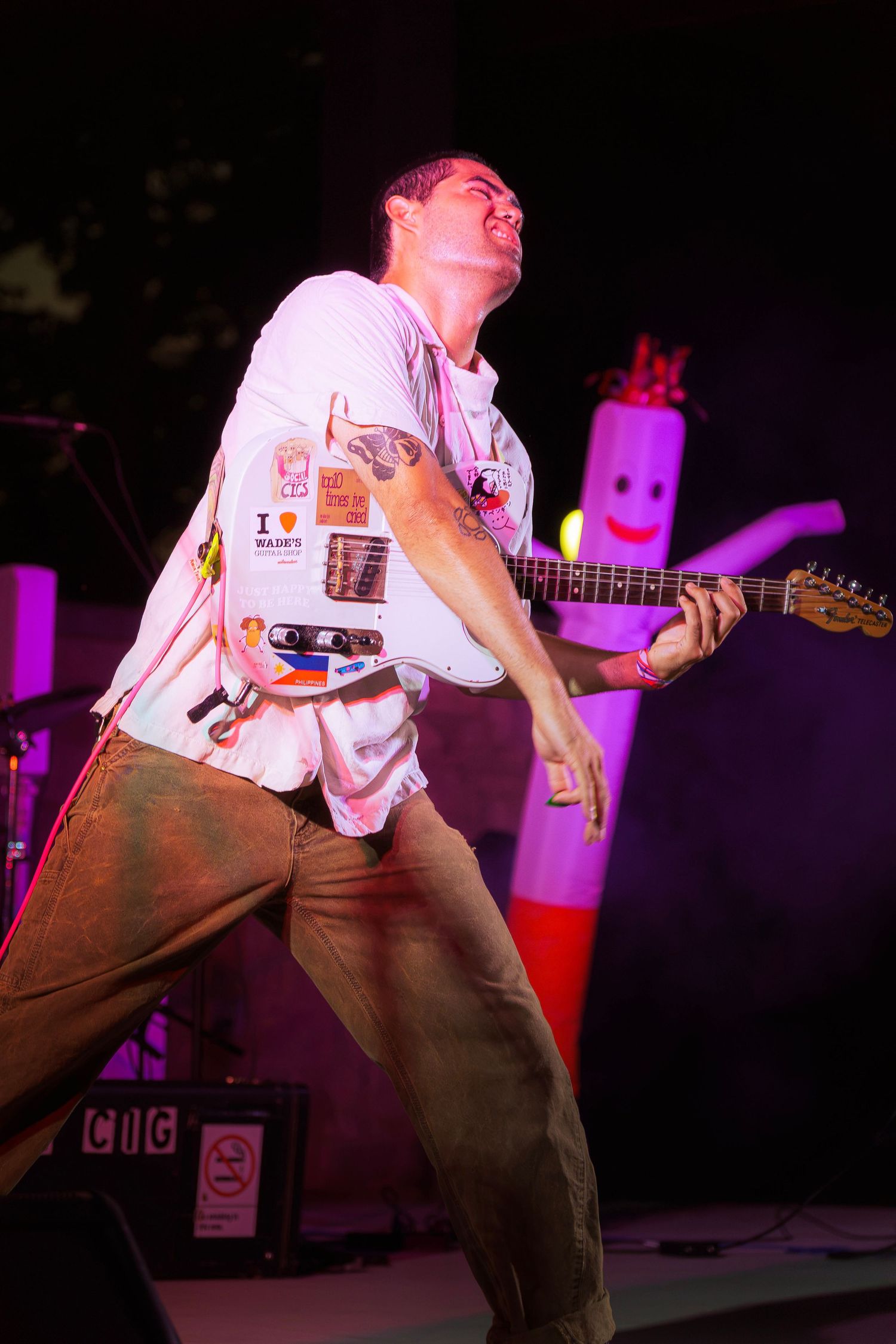 Guitarist performs energetically on stage, wearing a white sticker-covered t-shirt, with an inflatable tube man visible behind him.