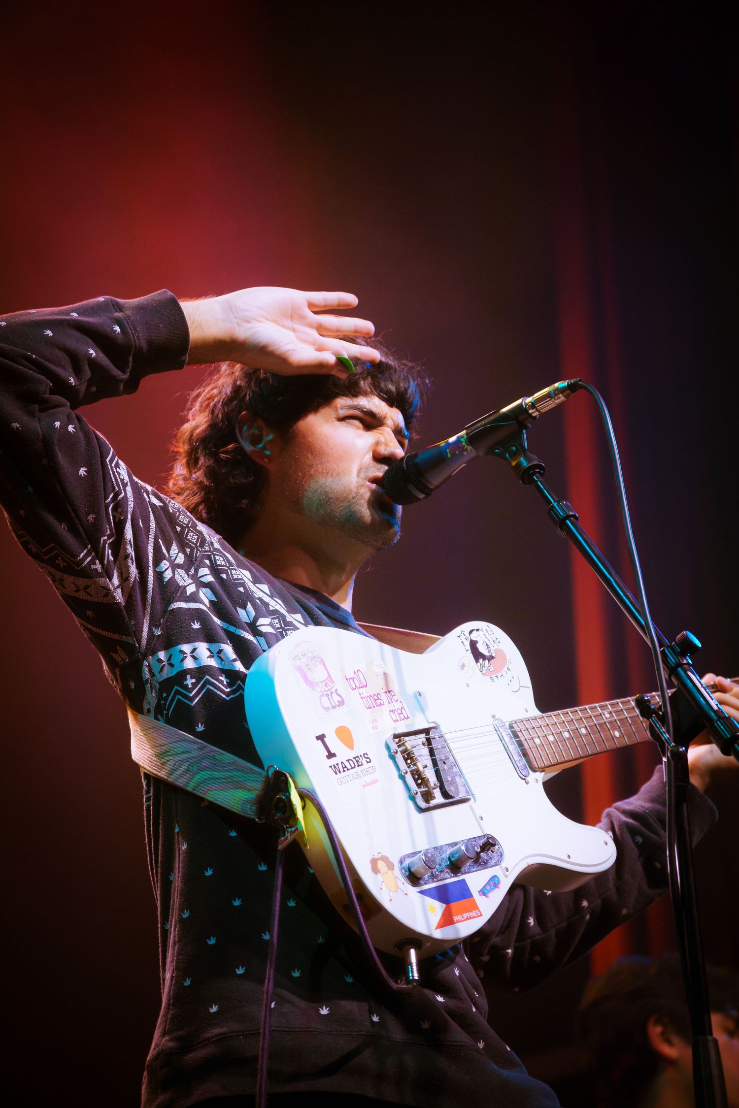 A performer in a dark cap sings into a microphone while playing a white electric guitar on stage with red lighting.